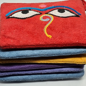 Op de afbeelding: Een stapel kleurrijke, vierkante, suède-achtige pouches. De bovenste pouch is rood met geborduurde ogen en een neus. Daaronder bevinden zich pouches in blauw, geel, paars en blauw. De pouches lijken handgemaakt.