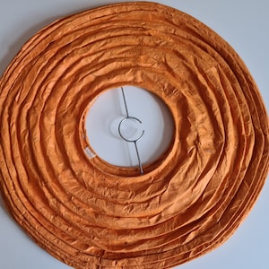 May include: Overhead view of an orange paper lantern shade. The shade is circular with concentric, accordion-like folds. A metal support structure is visible in the center. The lantern is designed to diffuse light.