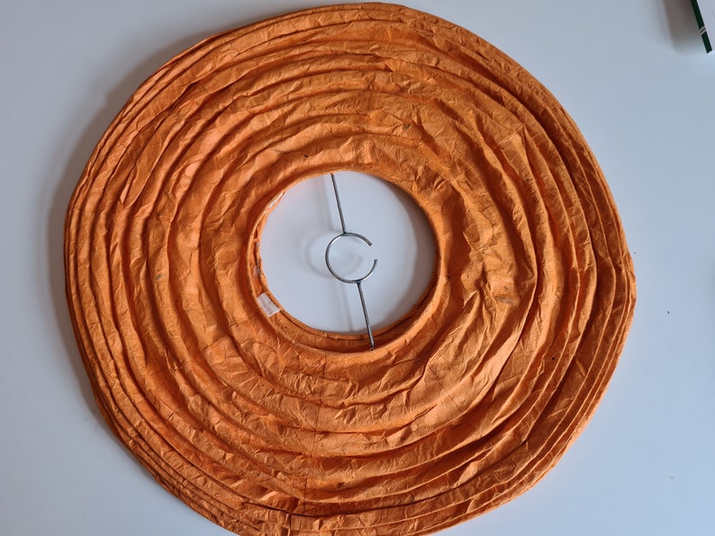 May include: Overhead view of an orange paper lantern shade. The shade is circular with concentric, accordion-like folds. A metal support structure is visible in the center. The lantern is designed to diffuse light.