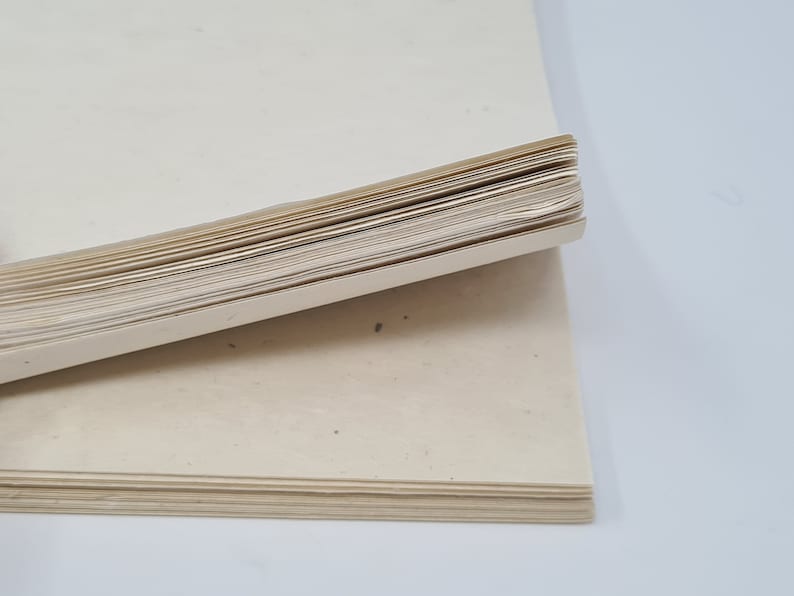 May include: A stack of blank, off-white paper sheets with a rough texture. The paper is folded in half and the edges are visible.
