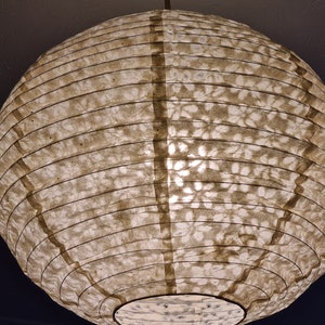 May include: A round, paper lantern with a warm, off-white hue. The lantern's surface has a textured, patterned design and is supported by dark, vertical bands. The light source is visible from within, creating a soft glow.