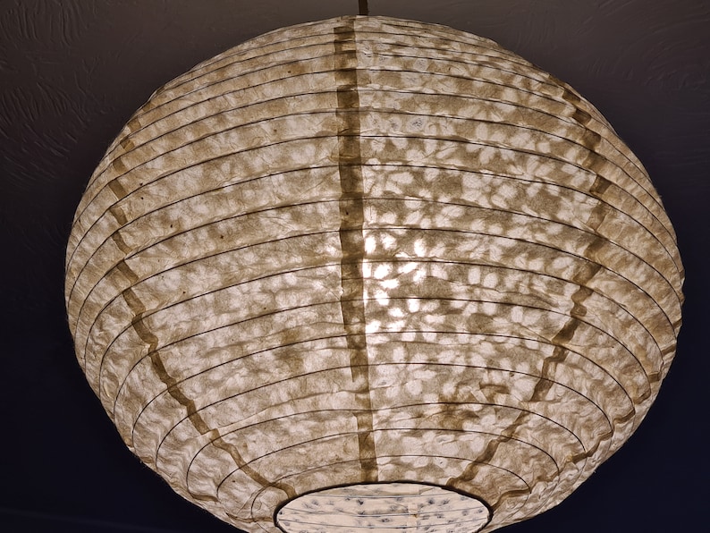 May include: A round, paper lantern with a warm, off-white hue. The lantern's surface has a textured, patterned design and is supported by dark, vertical bands. The light source is visible from within, creating a soft glow.