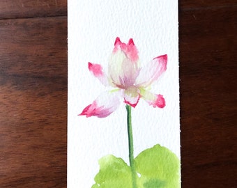 Puede incluir: Una pintura de acuarela de una flor de loto rosa y blanca con hojas verdes sobre un fondo blanco. La pintura está en un marcapáginas con una cinta verde.