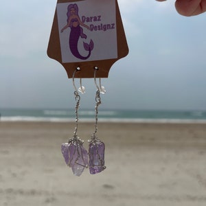 Könnte beinhalten: Ein Paar Ohrringe mit Amethyst-Kristallen, die in Silberdraht gewickelt sind. Die Ohrringe hängen an einer braunen Karte mit einer Meerjungfrauen-Illustration und dem Text "Daraz Designz".