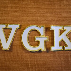 Op de afbeelding: Witte letters "VGK" met gouden rand, tegen een achtergrond van houtnerf. De letters zijn dik en blokkerig, met een getextureerde uitstraling. De gouden rand voegt een vleugje elegantie toe aan het ontwerp.