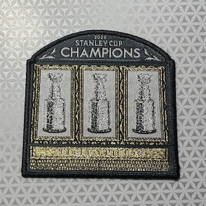 Può includere: Toppa ricamata con il design della Stanley Cup. La toppa è nera con finiture dorate e le parole "2023 STANLEY CUP CHAMPIONS" in alto. Sotto ci sono tre immagini della Stanley Cup. Le parole "GOLDEN KNIGHTS" sono in basso.