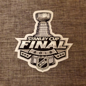 Op de afbeelding: Een witte en zilveren geborduurde patch met een Stanley Cup Final-logo. Het logo toont een hockeystick, een puck met het NHL-logo en de tekst "Stanley Cup Final 2018".