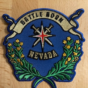 Può includere: Una toppa ricamata blu e oro con una stella e spade incrociate, circondata da una ghirlanda di foglie verdi e fiori gialli. Il testo "BATTLE BORN" è su uno stendardo dorato sopra la stella e "NEVADA" è sotto la stella.