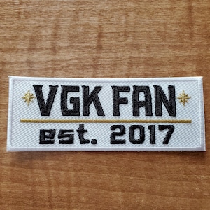 May include: White embroidered patch with black text that reads "VGK FAN" with a gold line and the text "Est. 2017" below.