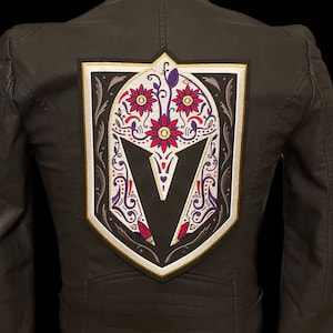 Può includere: Giacca di pelle nera con una grande toppa ricamata sul retro. La toppa presenta un teschio bianco con una 'V' stilizzata al centro, circondata da motivi floreali in rosso, viola e giallo.