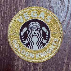 May include: A round, gold glitter patch with the words "VEGAS GOLDEN KNIGHTS" in white lettering. The center features a black and white logo of a woman with hockey sticks. The patch is on a dark wood surface.