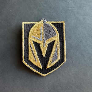 May include: A black, gold, and gray embroidered patch featuring a knight's helmet with a V shape on the front.