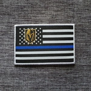 Könnte beinhalten: Ein schwarz-weißes amerikanisches Flaggen-Patch mit einer blauen Linie in der Mitte. Die Flagge hat ein goldenes und weißes Logo eines Ritterhelms in der oberen linken Ecke.