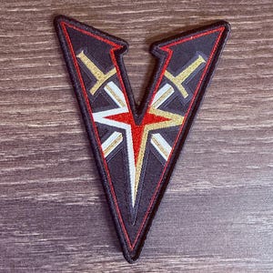 May include: A V-shaped embroidered patch featuring a black background with a red and gold star design at the center. Two gold swords are crossed behind the star, and the entire emblem is outlined with a thin red border and black stitching.