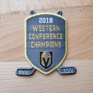 Può includere: Una toppa ricamata grigia e dorata con il testo "2018 Western Conference Champions" e un logo "V" stilizzato. La toppa ha la forma di uno scudo e due bastoni da hockey incrociati.