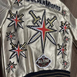 Peut inclure: Une veste en cuir dor&eacute;e avec un col et des poignets ray&eacute;s noir et or. La veste comporte plusieurs patchs, dont une grande &eacute;toile avec des &eacute;p&eacute;es, un patch qui dit "Golden Misfits" et un patch qui dit "All-Star Las Vegas 2022".