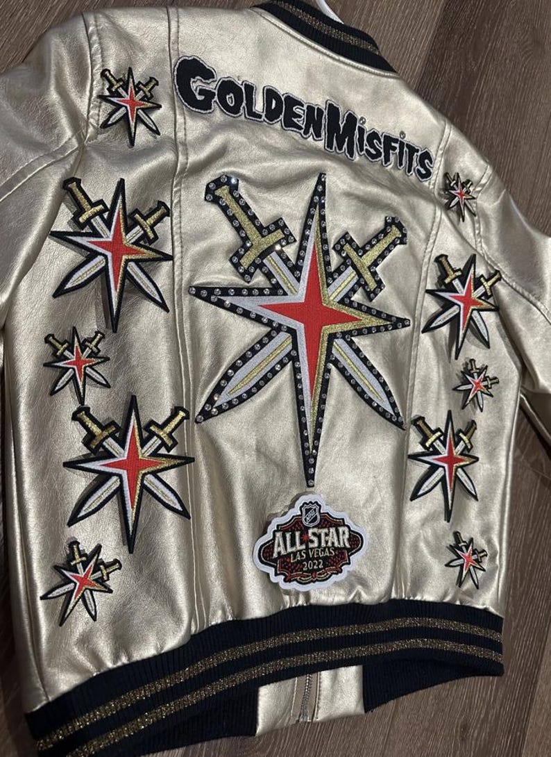 Peut inclure: Une veste en cuir dor&eacute;e avec un col et des poignets ray&eacute;s noir et or. La veste comporte plusieurs patchs, dont une grande &eacute;toile avec des &eacute;p&eacute;es, un patch qui dit "Golden Misfits" et un patch qui dit "All-Star Las Vegas 2022".