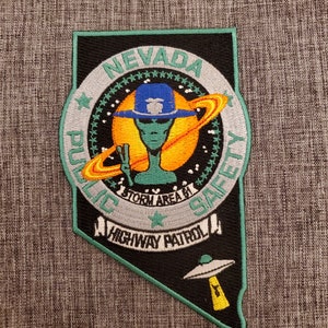 May include: A green, black, and white embroidered patch with the words "Nevada Public Safety Highway Patrol" and a cartoon alien wearing a police hat and holding a peace sign. The alien is in front of a yellow sun with a ring around it.