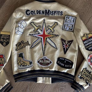 Peut inclure: Une veste bomber dor&eacute;e avec divers patchs, dont le logo des Golden Knights, le logo de Vegas et le patch Western Conference Champions 2022-23. La veste pr&eacute;sente &eacute;galement une &eacute;toile avec des &eacute;p&eacute;es et le texte "Golden Misfits".