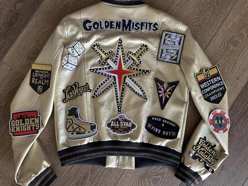 Peut inclure: Une veste bomber dor&eacute;e avec divers patchs, dont le logo des Golden Knights, le logo de Vegas et le patch Western Conference Champions 2022-23. La veste pr&eacute;sente &eacute;galement une &eacute;toile avec des &eacute;p&eacute;es et le texte "Golden Misfits".