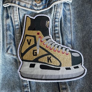 May include: A gold, black, and gray embroidered patch in the shape of a hockey skate. The patch features the letters "V", "G", and "K" on the skate. The patch is sewn onto a light blue denim jacket.