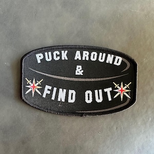 May include: Black and white embroidered patch with a hockey puck shape. The text "Puck Around & Find Out" is written in white with a red star on each side.