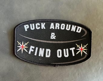 May include: Black and white embroidered patch in the shape of a hockey puck. The text "Puck Around & Find Out" is written in white with a red star on each side.