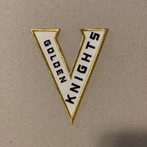 May include: A white felt patch with a gold outline in the shape of a V. The words "GOLDEN KNIGHTS" are written in black inside the V.