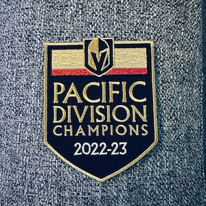 May include: A black and gold embroidered patch with the text "Pacific Division Champions 2022-23". The patch features a stylized hockey helmet with a "V" inside.