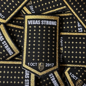 May include: A close-up of several black and gold patches with the text "VEGAS STRONG" and "1 OCT 2017". The patches are made of fabric and have a glittery finish.
