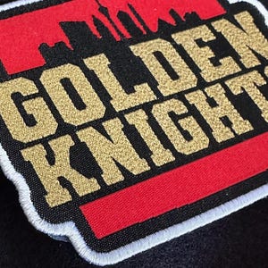 Può includere: Toppa ricamata con la scritta "GOLDEN KNIGHTS" in filo dorato su sfondo nero. Un rettangolo rosso con uno skyline della città è sopra il testo. La toppa ha un bordo bianco, probabilmente per l'abbigliamento.