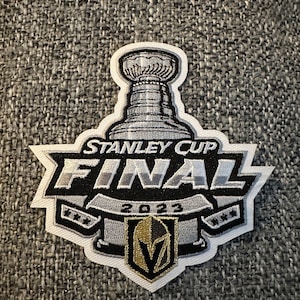 May include: A white patch with a black and silver Stanley Cup Final logo. The logo features a silver Stanley Cup trophy, the words "Stanley Cup Final" and the year "2023". A gold and black Vegas Golden Knights logo is below the banner.