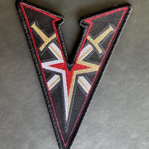 May include: A black and white embroidered patch in the shape of a V with a red and gold design. The patch has a red border and is embroidered with two swords.