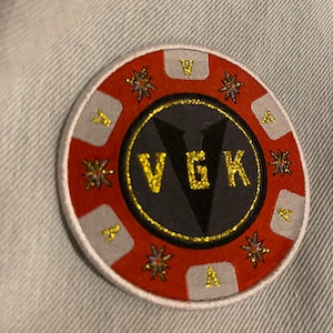 May include: A round, embroidered patch with a red and white border. The center features the letters "VGK" in gold against a black background. Gold accents and starburst designs are also present.