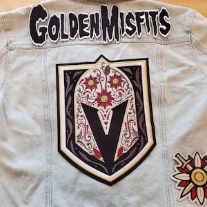 May include: A light-wash denim jacket featuring a large back patch with a shield design containing a stylized helmet, floral patterns, and a central V. Above the shield is a patch reading GOLDEN MISFITS, and a smaller floral patch with crossed swords.