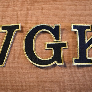 Può includere: Tre lettere in feltro nero con cuciture dorate, che compongono la parola "VGK".