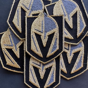 Puede incluir: Un conjunto de seis parches bordados en negro y dorado con un diseño de casco estilizado. El casco está bordeado de dorado y tiene un diseño a rayas negro y dorado.