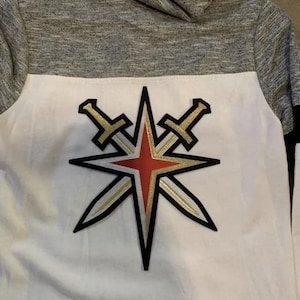 Può includere: Felpa con cappuccio bianca e grigia con un logo a forma di stella rossa, oro e nera con due spade incrociate dietro. Il logo è centrato sul davanti della felpa.