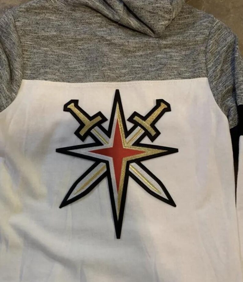 Peut inclure: Sweat-shirt &agrave; capuche blanc et gris avec un logo d'&eacute;toile rouge, or et noir avec deux &eacute;p&eacute;es crois&eacute;es derri&egrave;re. Le logo est centr&eacute; sur le devant du sweat-shirt.