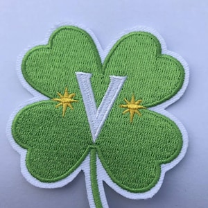 Può includere: Un cerotto a forma di trifoglio a quattro foglie verde con una lettera V bianca al centro e due stelle gialle su entrambi i lati della lettera.