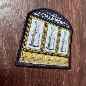 Puede incluir: Un parche negro, dorado y blanco que conmemora a los Campeones de la Copa Stanley 2023. El parche presenta tres imágenes del trofeo de la Copa Stanley y las palabras "Champions" y "Golden Knights".