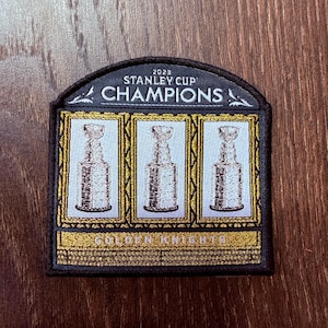 May include: A black and gold patch commemorating the 2023 Stanley Cup Champions. The patch features three Stanley Cup trophies in gold frames, with the words "STANLEY CUP CHAMPIONS" and "GOLDEN KNIGHTS".