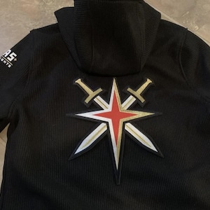 Può includere: Giacca con cappuccio nera con il logo dei Vegas Golden Knights sul retro. Il logo presenta una stella rossa con dettagli oro e bianchi e due spade incrociate. Il nome della squadra è stampato sulla manica.