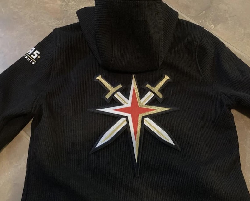 Peut inclure: Sweat-shirt &agrave; capuche noir avec un grand patch brod&eacute; au dos. Le patch repr&eacute;sente une &eacute;toile rouge avec deux &eacute;p&eacute;es crois&eacute;es, le tout en or. Le texte "LAS" est visible sur la manche gauche.