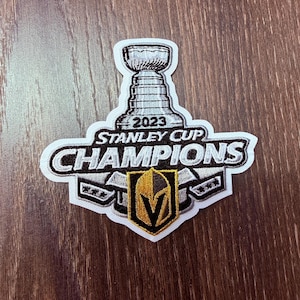 May include: Embroidered patch featuring the Stanley Cup trophy and the text "2023 Stanley Cup Champions." The patch has a white border, with a gold and black emblem at the bottom. The patch is on a wooden surface.