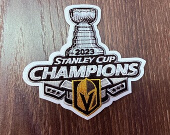 Peut inclure: Écusson brodé avec le trophée de la Coupe Stanley et le texte "2023 Stanley Cup Champions". L'écusson a une bordure blanche, avec un emblème doré et noir en bas. L'écusson est sur une surface en bois.