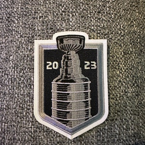 Può includere: Una toppa ricamata in argento e nero con un trofeo di hockey con i numeri 20 e 23 su entrambi i lati.