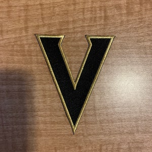 May include: A black felt letter V with a gold outline. The letter is on a wood background.