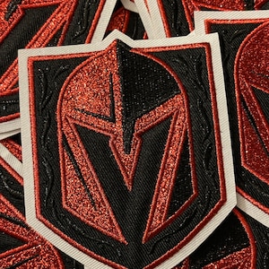 Può includere: Un mucchio di toppe ricamate rosse e nere che raffigurano un elmo da cavaliere con un designo rosso e nero. Le toppe sono bordate con cuciture bianche.