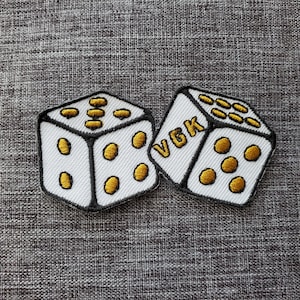 May include: Two white dice patches with black borders and gold-colored dots. One die has the letters "VGK" in gold. The dice are embroidered and appear to be made of fabric. The background is a textured gray surface.